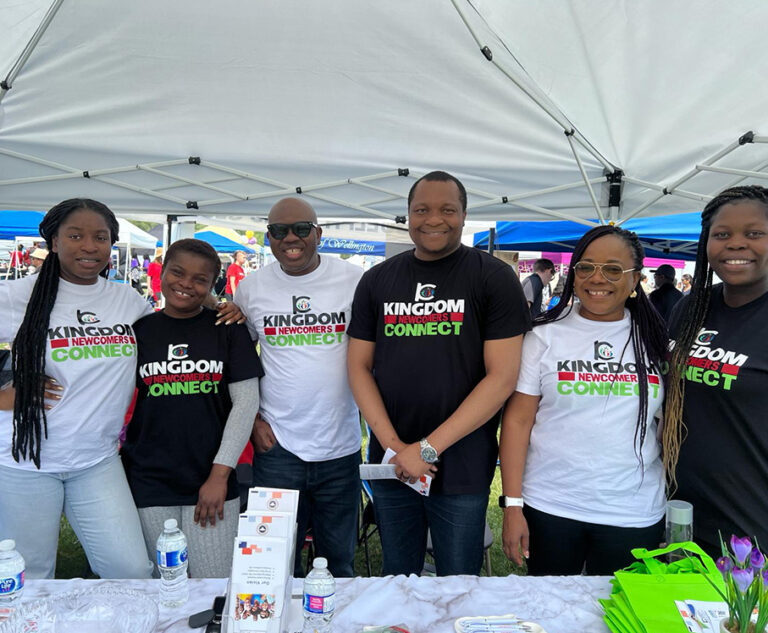 Six team members in Kingdom Newcomers Connect T-shirts smile at their booth at an outdoor community event, with brochures and water bottles on the table.